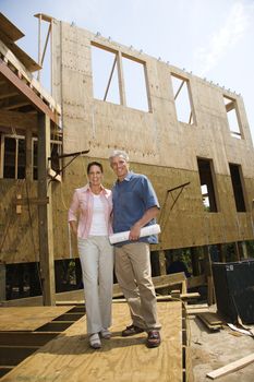 Caucasian mid-adult male holding blue prints with arm around mid-adult female in building construction site.