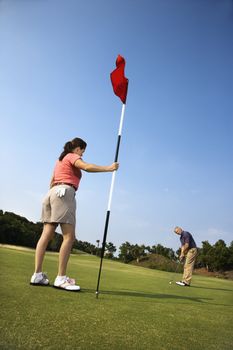 Caucasion mid-adult man putting golfball while Caucasion mid-adult woman holds flag.