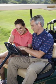 Mid-adult male and female in golf cart pointing at score card.