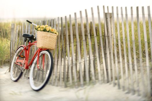 Red vintage bicycle with basket and flowers leaning against wooden fence at beach.