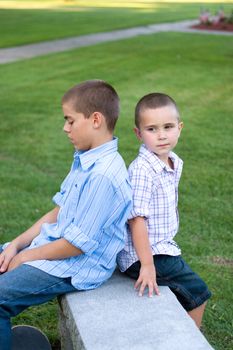 Two brothers sitting back to back on a bench in the park.  