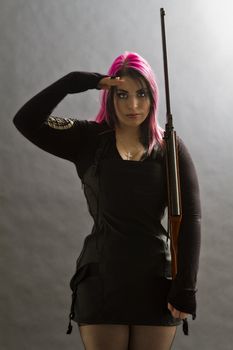 Girl wearing goth clothes with pink and black hair doing a military salute and holding a rifle