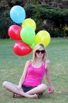 Beautiful young blond lady sitting on green grass with colorful balloons