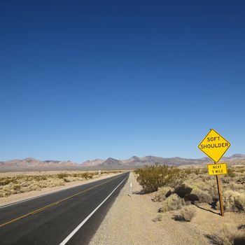 Road in desert with sign for soft shoulder and mountains in distance.