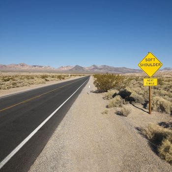 Road in desert with sign for soft shoulder and mountains in distance.