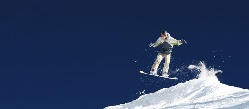 A snowboard jumps through the air in front of a crisp sky background. The snow spray has great detail. Lots of copy space