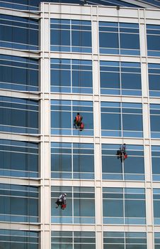 Three window washers descend on ropes high above the city. The building is a very modern glass structure.