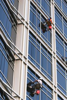 Two window washers descend on ropes high above the city. The building is a very modern glass structure.