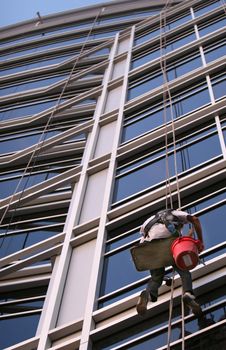 A window washer descend on ropes high above the city. The building is a very modern glass structure.