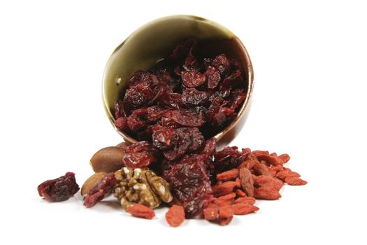 Red ripe dried cranberries spilling from a small bowl with mixed nuts and goji berries on a reflective white background