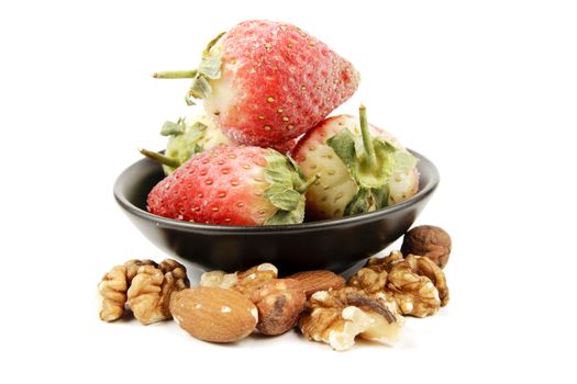 Red ripe frozen strawberries in a small black bowl with mixed nuts on a reflective white background