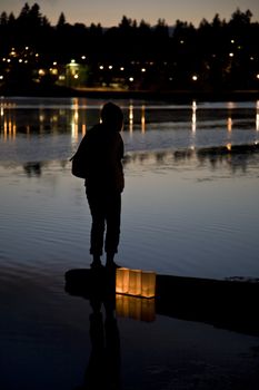 A silhouetted figure stands on a log protruding into a lake seemingly oblivious to the three candlelit lanterns behind them
