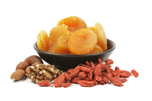 Dried juicy orange apricots with mixed nuts and goji berries in a small black bowl on a reflective white background