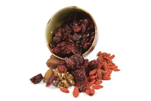 Red ripe dried cranberries spilling from a small bowl with mixed nuts and goji berries on a reflective white background