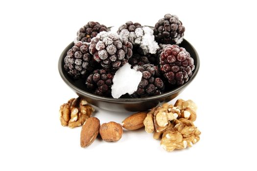 Ripe frozen blackberries in a small black bowl with mixed nuts on a reflective white background