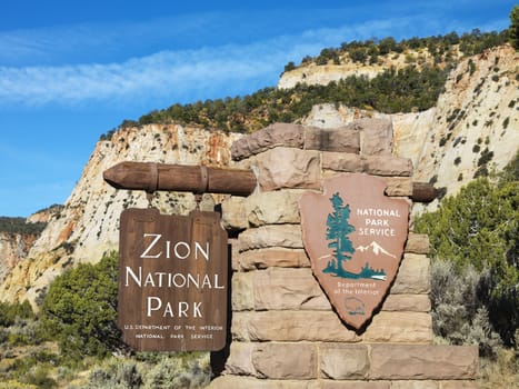 Wooden and stone sign for Zion National Park, Utah with rocky cliffs in the background.