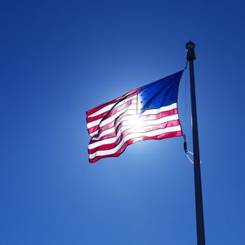 American flag on flagpole waving against blue sky with sun shining through from behind.