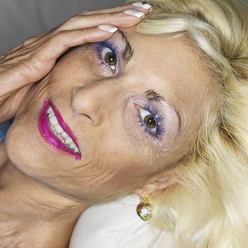 Portrait of tan Caucasion blonde middle-aged woman wearing lots of makeup with hand on face smiling at viewer.