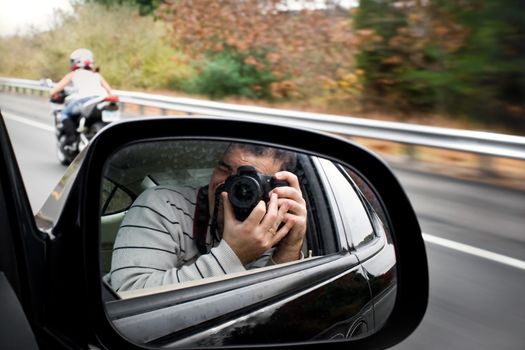 A paparazzi photographer takes a photo of a woman driving a motorcycle on the highway. Shallow depth of field.
