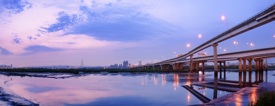 Panoramic cityscape with modern building and bridge under dramatic sky and illuminated reflection on sand and water of river in Taipei, Taiwan.