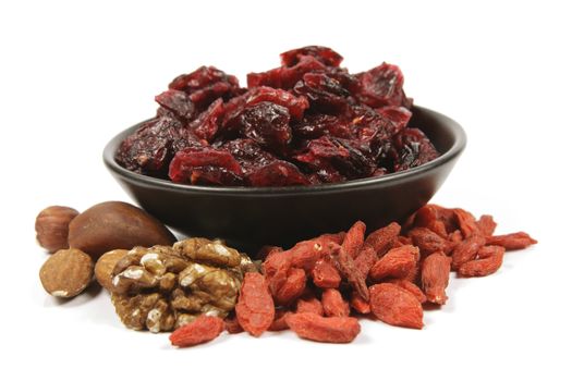 Red ripe dried cranberries in a small black bowl with mixed nuts and goji berries on a reflective white background