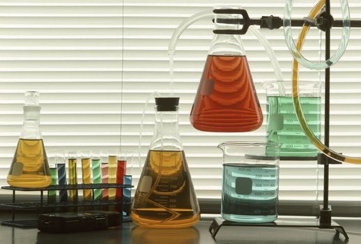 Scientific glassware and tubes filled with colored liquids against window blinds