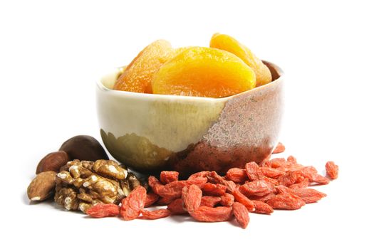 Dried juicy orange apricots in a green and brown bowl with mixed nuts and goji berries on a reflective white background
