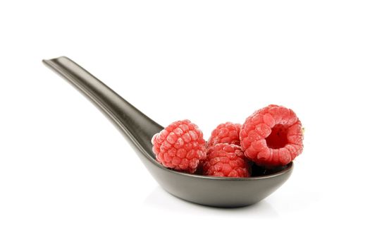 Red ripe frozen raspberries on a small black spoon with a reflective white background