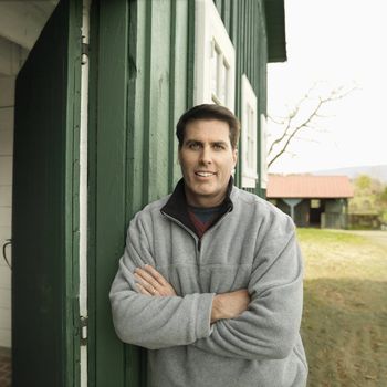 Caucasian man leaning against building smiling in rural setting.