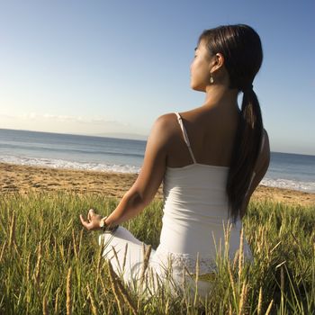 Young Asian female sitting on beach meditating and looking out to ocean.