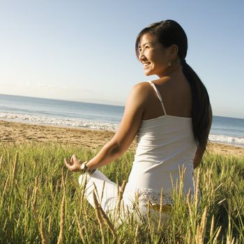 Young Asian female sitting on beach meditating and looking off to the side.