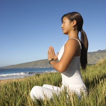 Young Asian female sitting on beach looking out to ocean with hands pressed together.