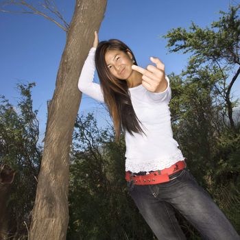 Young adult Asian female with one hand on tree and other hand giving the viewer the middle finger.