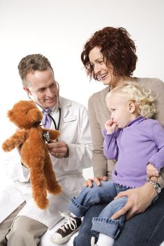 Middle-aged adult Caucasian male doctor holding stethoscope to teddy bear while Caucasian mother and daughter watch.