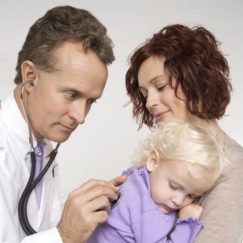 Middle-aged adult Caucasian male doctor holding stethoscope to female toddler's back with mother watching.