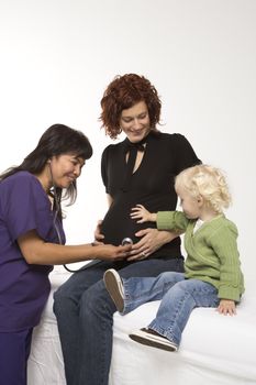 Nurse holding stethoscope on Caucasian pregnant woman's belly as daughter holds hand on belly.