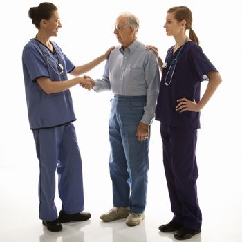 Mid-adult Caucasian female in scrubs shaking hand of elderly Caucasian male with another mid-adult Caucasian female with hand on his shoulder.