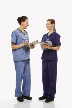 Portrait of Caucasian women doctors in medical scrubs standing talking holding medical charts against white background.