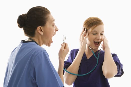 Portrait of Caucasian healthcare workers wearing scrubs with one yelling into stethoscope that is attached to other's ears against white background.