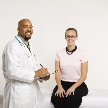 Mid-adult African-American doctor with clipboard and Caucasion mid-adult female patient in doctor's office looking at viewer and smiling.