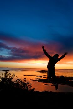 A person jumping by the ocean raising their arms in celebration with slight motion blur on the hands.