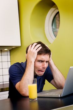 A worried computer user sits in his kitchen with a desperate look on his face