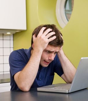 A young male at home in the kitchen looking worried while using a computer.