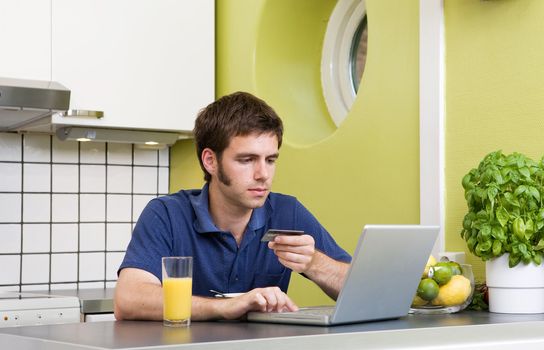 A young male shops online with a credit card in his kitchen.