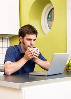 A young man uses the computer in the kitchen while enjoying a warm drink.