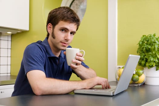 A young man uses the computer in the kitchen while enjoying a warm drink