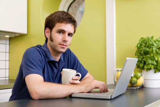 A young man uses the computer in the kitchen while enjoying a warm drink