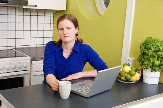 A young woman sitting in the kitchen with a coffee and a computer looking at the camera.