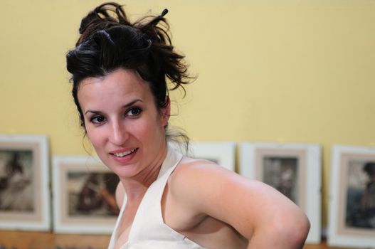 A young woman with dark hair leans on one arm while glancing to the right, a yellow wall and picture frames in the background.