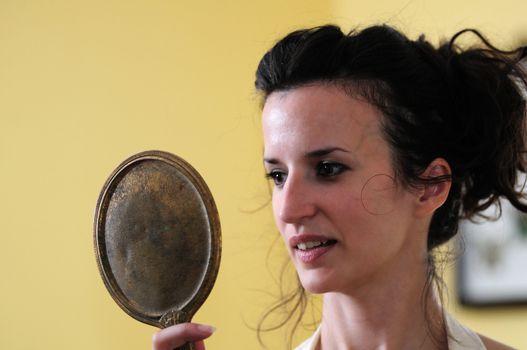 A young woman gazes at her reflection in an antique hand mirror against the background of a yellow wall.